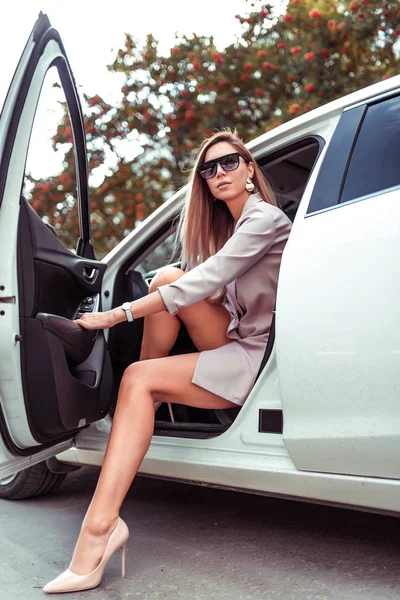 stylish fashionable beautiful Woman enters from interior of white car, business class sedan. Taxi, car sharing, summer city. Tanned figure sunglasses pink suit. parking lot, trees road background.