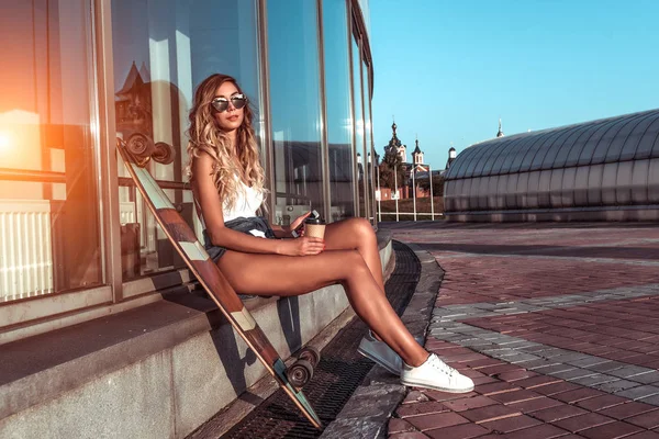 Schöne stilvolle modische Mädchen Sommer Stadt sitzt Fenster Gebäude ruht Sonnenbaden Badeanzug, trägt eine Brille mit Longboard-Skateboard. freier und offener Raum für Text. Jugendurlaub im modernen Stil. — Stockfoto