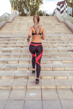 Güzel bir kız sabah antrenmanı merdivenlerde, yazın şehir, arkadan manzara. Spor eğitimi aktif yaşam tarzı. Bronz tenli atletik kadın. Spor Giyim Kıyafeti Üstü. Uzun saçlı ince bir figür..