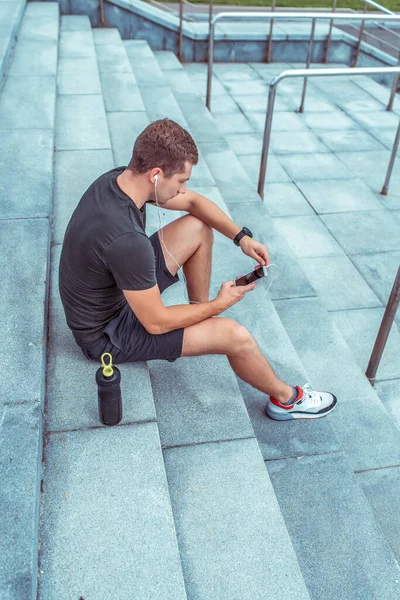 A man is resting after a workout, in the summer in the city, sportswear, mobile phone headphones, listening to music in the application. Bottle shaker with protein. Sits on the steps. — ストック写真