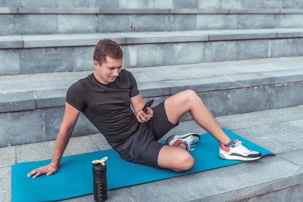 Man is resting after training on mat, in summer in city, sportswear, mobile phone. Bottle shaker with protein. Sits on steps. Reads a message, sending SMS on social networks. — ストック写真