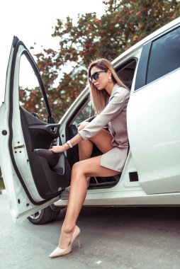 Güzel kız beyaz iş arabası sedanından iniyor. Yazın şehirde, otoparkta. Şık takım elbise, uzun saç. Yüksek topuklu ayakkabılar. Araba parkı. Modern bir kadının bronzlaşmış figürü. Kapı açık..