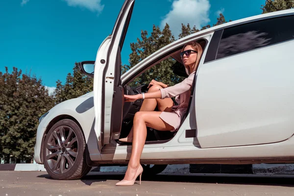 Mulher dirigindo um carro, no verão no estacionamento sai e entra no carro. Uma mulher de negócios de fato e sapatos. Óculos de sol, negócio branco VIP táxi sedan, aluguel de carros, compartilhamento de carros . — Fotografia de Stock