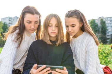 Üç liseli kız öğrenci 13-15 yaşlarında, yaz günü şehir videoları izliyor akıllı telefon, online uygulama sosyal ağları internet. Sıkı örülmüş kazaklar. En iyi arkadaşlar okuldan sonra dinlenir..