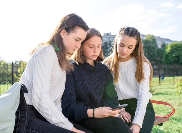 3 dívky přítelkyně teenageři 14-15 let, sedět lavička podzim odpoledne park relaxovat po vyučování na vysoké škole, sledovat videa sociální sítě smartphone obrazovce. V neformálním oblečení, svetru a džínách. — Stock fotografie