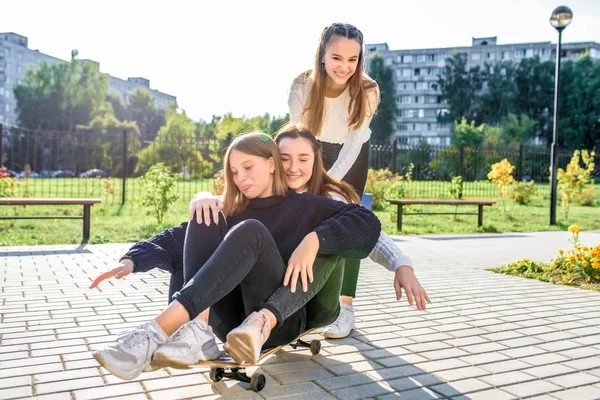 3 niñas colegialas adolescentes 13-15 años, en el día de otoño, ciudad de verano, montar un monopatín, sonrisas felices, regocijarse y reír. Descansa en la calle después de la escuela. Suéter vaquero casual . —  Fotos de Stock