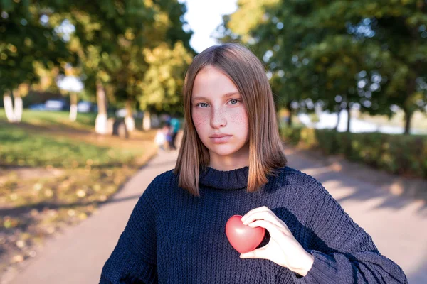 Девочка-подросток 12-15 лет, летом в городе, держит в руке игрушечное сердце, концепт-подарок любви родительской помощи и поддержки. Тёплый вязаный свитер . — стоковое фото