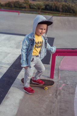 3-5 yaşlarında küçük bir çocuk, kaykaya biniyor, hafta sonu dinleniyor, eğitim ve antrenman, tahtadaki ilk adımlar. Yazın, şehirde. Günlük kıyafetler, kapüşonlu kot pantolon..