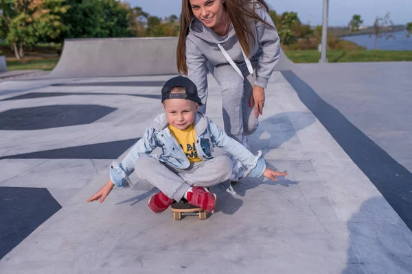 Anak laki-laki berusia 3-5 tahun, ibu wanita, tersenyum bahagia, bahagia, belajar mengendarai skateboard, di musim panas di taman kota. Istirahat akhir pekan di lapangan olahraga. Pakai casual skateboard . — Stok Foto