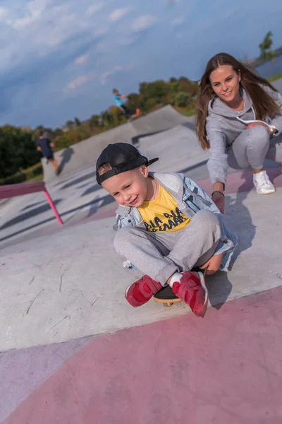 Anak laki-laki berusia 3-5 tahun, ibu perempuan, pelatih bersenang-senang, tersenyum, bersukacita, belajar mengendarai skateboard di musim panas di taman kota. Istirahat akhir pekan di lapangan olahraga. Pakai casual skateboard . — Stok Foto