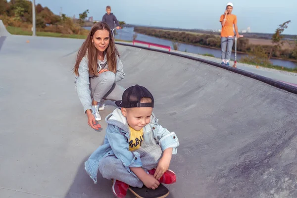 Anak laki-laki berusia 3-5 tahun, ibu wanita, senang bermain dan bersenang-senang, belajar mengendarai skateboard, di musim panas di taman kota. Istirahat akhir pekan di lapangan olahraga. Pakai casual skateboard . — Stok Foto