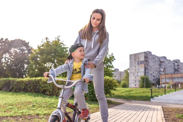 Happy ceria keluarga muda, anak kecil 3-5 tahun, ibu wanita, belajar untuk naik sepeda, di musim panas di taman kota. Istirahat akhir pekan di lapangan olahraga. Pakaian kasual. Emosi kasih sayang dan pengasuhan . — Stok Foto