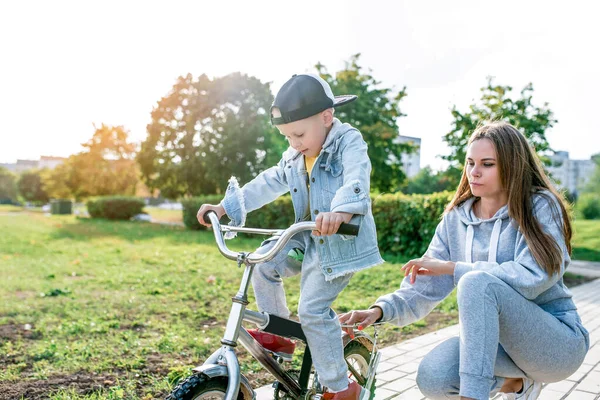 Keluarga muda, anak laki-laki berusia 3-5 tahun, ibu wanita, belajar mengendarai sepeda, di musim panas di taman kota. Istirahat akhir pekan di lapangan olahraga. Pakaian kasual. Emosi kasih sayang dan pengasuhan . — Stok Foto