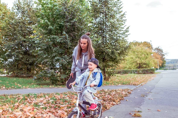 Νέα οικογένεια, μικρό αγόρι 3-5 ετών, μαμά γυναίκα, διδάσκονται να οδηγούν ένα ποδήλατο, εκπαίδευση βοήθεια και υποστήριξη σε μαθήματα ισορροπίας. Το καλοκαίρι στην πόλη. Χαλαρά ρούχα, τζιν. Ελεύθερος χώρος για κείμενο αντιγραφής. — Φωτογραφία Αρχείου