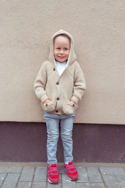 Malý chlapec 3-5 let, šťastně se usmívá a stojí u zdi. Nenucené teplé oblečení s kapucí. Podzimní den. Čekání na rodiče a přátele. — Stock fotografie