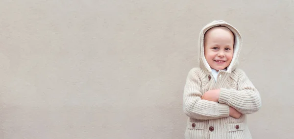 Malý chlapec 3-5 let, šťastný úsměv a radost, béžová zeď pozadí. Nenucené teplé oblečení s kapucí. Podzimní den. Volné místo pro kopírování textu. Emoce štěstí a potěšení. — Stock fotografie