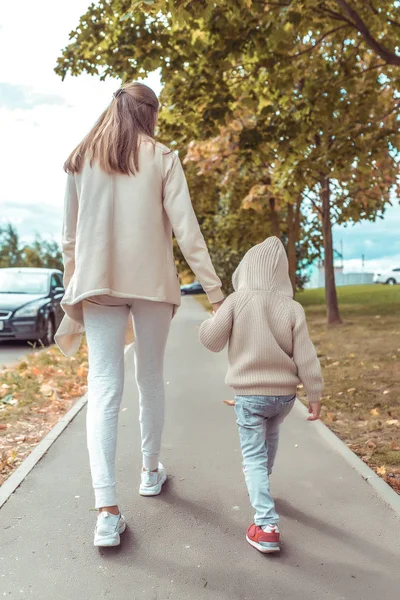 Ibu wanita berjalan dengan anaknya, anak laki-laki berusia 4-5 tahun, hari musim gugur di jalan di kota, jalan latar belakang dan pohon-pohon, pakaian hangat kasual dengan tudung . — Stok Foto