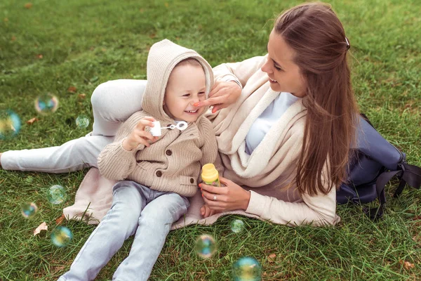 Žena matka s dítětem, syn chlapec 4-5 let, podzim na trávníku, foukání mýdlové bubliny dovolená, emoce štěstí, radost smích potěšení, ležérní teplé oblečení. Rodičovské děti. — Stock fotografie