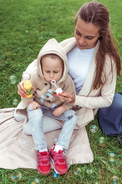 Женщина мать с ребенком, маленький сын мальчик 4-5 лет, осень на газоне, дующие мыльные пузыри каникулы, эмоции счастья, радость ухода за детьми, случайная теплая одежда. Родительские дети . — стоковое фото