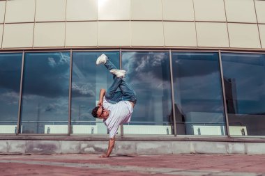 Tek kollu bir adam, genç bir dansçı, yazın şehirde dans ediyor, sokak dansları yapıyor, modern bir gençlik tarzı. Moda ve fitness sporu. Metni kopyalamak için boş alan. Cam pencere bulutları arkaplan.