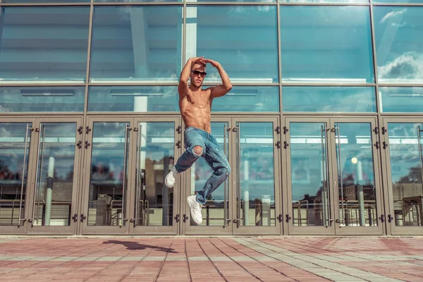 Jumping man, muscular torso, young guy dancer, in summer in the city, dancing street dances, modern youth style. Fashion and fitness sport. Free space for copy text. Background glass doors. — Stock Photo, Image