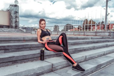 Kız sporcu, yaz şehri, idmandan sonra dinleniyor, merdivenlerde oturuyor, akıllı telefondan mesaj yazıyor, kulaklıkla müzik dinliyor. Zevk ve keyif motivasyonu duyguları..