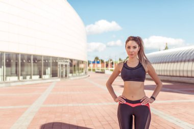 Güzel ve atletik, bronz tenli bir kız, yazın şehirde, fitness antrenmanında, önden poz veriyor, spor kıyafetleri üstte. Binanın arka planı. Metin motivasyonu için boş alan.
