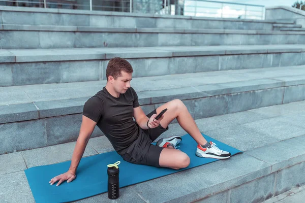 Männliche Sportler, in der Sommerstadt, nach dem Training ausruhen, ein Smartphone in der Hand, Nachrichten lesen und schreiben auf dem Smartphone, sich auf einer Fitnessmatte ausruhen. Flaschenschüttler mit Eiweiß und Wasser. — Stockfoto