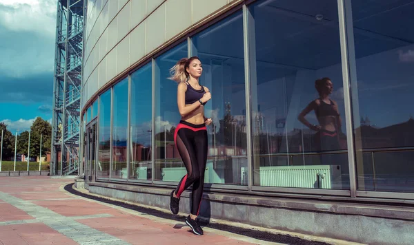 Menina bonita, verão na cidade, treinamento de fitness na rua, jogging de manhã, sportswear, leggings top. Uma figura bronzeada. Movimento em salto. Janelas de vidro de fundo do edifício. Espaço livre texto cópia . — Fotografia de Stock