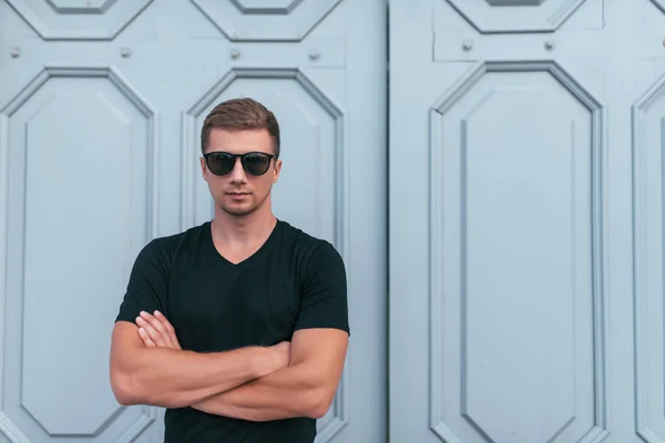 Homem em camiseta preta e óculos de sol, no verão no fundo da cidade de portas de madeira, posando, moda e estilo moderno, espaço livre para texto de cópia. Emoções de positivo e auto-confiança . — Fotografia de Stock