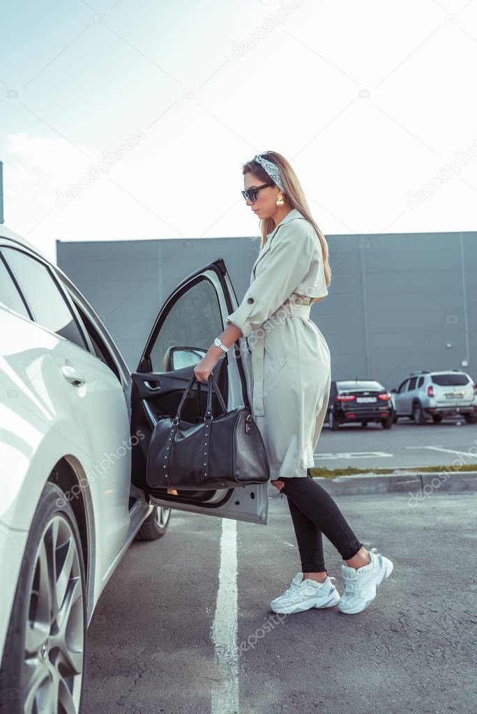 woman in summer in city opens car door, parking shopping center, grocery trip, modern clothes, a raincoat, black sneaker jeans, leather bag. Taxi call, car sharing.