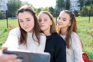 12-14 yaşlarındaki üç genç kız, yaz mevsiminde telefonda fotoğraf çekiyor, mutlu bir gülümseme pozu veriyor. Sosyal ağlar, internet, uygulama akıllı telefondan videolar izliyor. Günlük kıyafetler, kalın kazaklar..