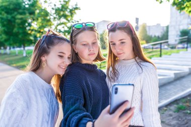 12-14 yaşlarında üç genç kız öğrenci, yaz şehri, el telefonu, mutlu bir şekilde gülümseyen, internetten fotoğraf izleyen. İnternetteki selfie fotoğrafı. Günlük kıyafetler, kalın kazaklar. Üniversiteye ara ver.