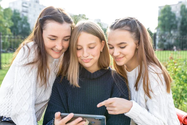 Три дівчини-підлітка віком 12-14 років, літнє місто дивляться відео-телефони, щасливі посміхаючись людям, які радіють. Соціальні мережі Інтернет, прикладний смартфон. Звичайний одяг, теплий светр.. — стокове фото