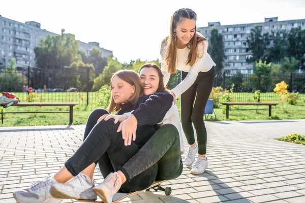 Drie tienermeisjes van 12-14 jaar oud, in de zomer in de stad, rijden op een skateboard, gelukkig glimlachen, plezier hebben en zich verheugen. Weekendje weg. Vrijetijdskleding, warme truien. Emoties van positieve en verrukking. — Stockfoto