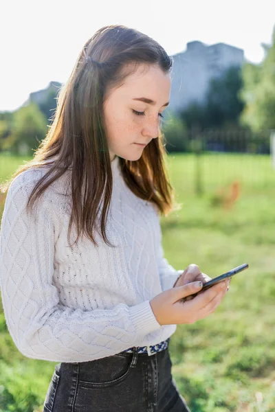 Tiener meisje van 12-15 jaar oud, houdt een mobiele telefoon in haar handen, in de zomer in het park, leest en schrijft een bericht in toepassing, sociale netwerken op internet. Casual wear, witte trui. — Stockfoto
