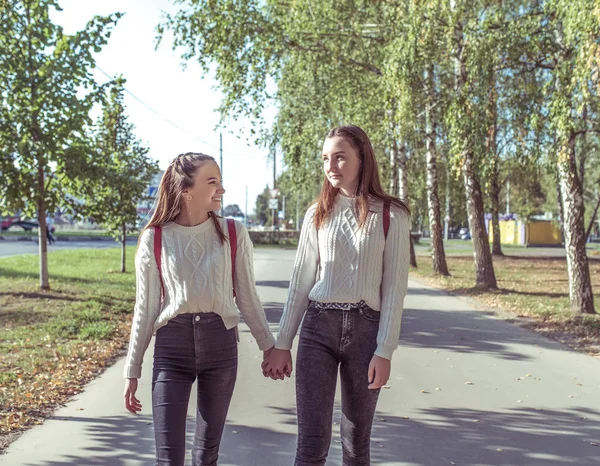 Due ragazze amiche studentesse 12-14 anni, passeggiata nel parco estivo, alberi stradali di sfondo, tenere per mano, ridere, divertirsi a parlare, tornare dopo la scuola, strada di casa. Emozioni di positivo e rilassante . — Foto Stock