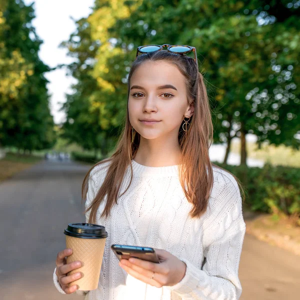 Gadis remaja berusia 10-12 tahun, memegang smartphone di tangannya, cangkir dengan teh kopi, musim panas di taman jalan, indah dan termenung. Liburan akhir pekan. Pakaian santai, rambut panjang, kacamata hitam. Pohon latar belakang . — Stok Foto