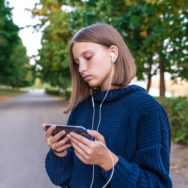 Tizenéves lány 12-15 éves, fogja a mobiltelefon kezét, nyáron a parkban, olvas és ír egy üzenetet alkalmazás, szociális hálózatok az interneten. Zenét hallgat fejhallgatón, — Stock Fotó
