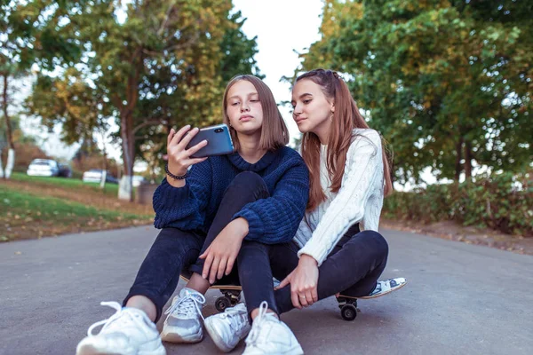 Two girls schoolgirls 12-14 years old, in summer in a city park, watch videos on a smartphone, sit on a skateboard, casual wear, a break in school and college, online official networks on Internet. — Stock Photo, Image