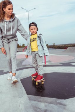 Kadın anne çocuğuna bakıyor, 4-5 yaşlarında mutlu bir çocuk, kaykay sürmeyi öğreniyor, yazın şehirde spor sahasında, kot şapkası giyerek. Rahatlama ve keyif duyguları..