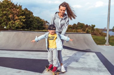 Kadın anne çocuğuna bakıyor, yardım öğreniyor, 4-5 yaşlarında küçük bir çocuk kaykay sürmeyi öğreniyor, yaz tatilinde spor sahasında kot şapkası takıyor. Rahatlama ve keyif duyguları..