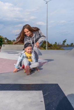Kadın anne çocuğuna bakıyor, yardım öğreniyor, 4-5 yaşlarında küçük bir çocuk kaykay sürmeyi öğreniyor, yaz tatilinde spor sahasında kot şapkası takıyor. Hafta sonu dinlen..