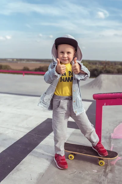 Šťastný malý chlapec 4-5 let, ukazuje palce nahoru gesto, učí se jezdit na skateboardu, v létě ve městě na sportovním hřišti, ležérní oblečení, džínová baseballová čepice. Emoce zábavy relaxace a požitku. — Stock fotografie