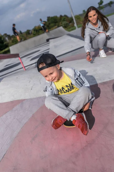 Woman mother teaches a child, help in balance, little boy 4-5 years old, learns to ride skateboard, in summer on sports ground in city, casual clothes, a denim baseball cap. Rest on the weekend. — Stock Photo, Image