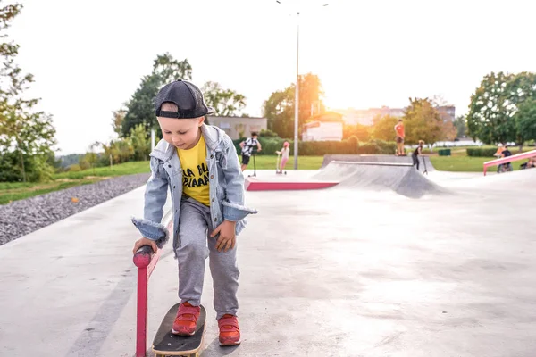 Anak kecil berusia 4-5 tahun, belajar mengendarai skateboard, di musim panas di kota di lapangan olahraga, memakai topi baseball denim. Istirahat di akhir pekan. Ruang kosong untuk menyalin teks . — Stok Foto