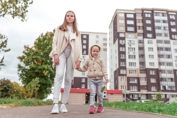 Junge Familie, Frau Mutter zu Fuß mit ihrem Sohn, Spaziergänge mit Kind, kleiner Junge 4-5 Jahre alt, Herbst in der Stadt, Gebäude Hintergrund Gehweg Asphalt. Freier Platz für Kopiertext. Lässig warme Kleidung. — Stockfoto