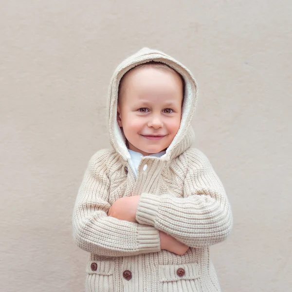 Маленький мальчик 4-5 лет, стоит бежевая стена в свитере с капюшоном, эмоциями счастья, радости, веселья и улыбки. Европейский ребенок в теплой одежде осенью и весной. вязанный свитер с капюшоном . — стоковое фото