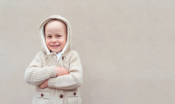 Malý chlapec 4-5 let, stojí béžová zeď, emoce štěstí, radosti, zábavy a úsměvu. Evropské dítě v teplých šatech na podzim a na jaře. Volné místo pro kopírování textu. Pletený svetr s kapucí. — Stock fotografie