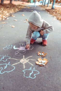 Küçük çocuk, yazın şehirde, pastel boyalarla resim çizer, günlük sıcak giysiler, kapüşonlu bej kazak. Yaratıcı yaratıcılık ve sanat konsepti. Arka planda sonbahar yaprakları asfalt. Metni kopyalamak için boş alan.